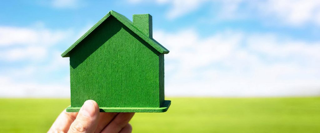 A green model of a house being held up against a backdrop of a green field with a cloudy blue sky.