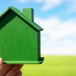 A green model of a house being held up against a backdrop of a green field with a cloudy blue sky.