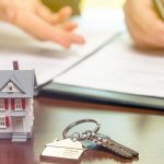 A real estate agent and customer sign contract papers with house keys and a small model home in front.