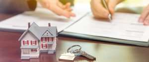 A real estate agent and customer sign contract papers with house keys and a small model home in front.