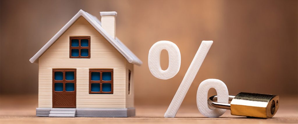 A decorative model house sits next to a large percent sign with a padlock attached symbolizing locking into a mortgage rate when buying a house.