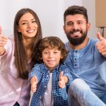 A family of three sitting between cardboard boxes and giving a thumbs up symbolizing the positive outlook of home buyers who have been waiting to buy a house.
