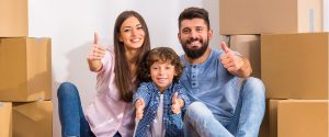 A family of three sitting between cardboard boxes and giving a thumbs up symbolizing the positive outlook of home buyers who have been waiting to buy a house.