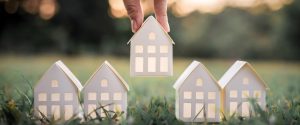 Miniature house models on grass with the center being lifted by a hand symbolizing the spring homes selling market.