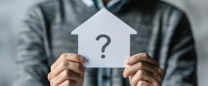 A man holding a paper cutout shaped like a house with a question mark inside it symbolizing the decision to buy a home.