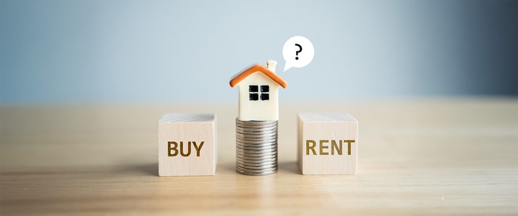 Wooden blocks labeled BUY and RENT on either side of a stack of coins topped by a small house model with a speech bubble containing a question mark illustrating the decision between renting vs buying a house.