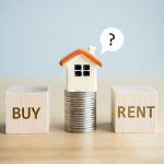 Wooden blocks labeled BUY and RENT on either side of a stack of coins topped by a small house model with a speech bubble containing a question mark illustrating the decision between renting vs buying a house.