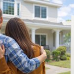 Couple standing arm in arm outside a modern suburban home at sunset, symbolizing the decision to buy a house in today’s real estate market.