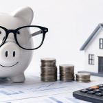 A piggy bank with glasses, a model home, coins, and a calculator resting on financial charts, representing housing affordability and saving for a home budget.