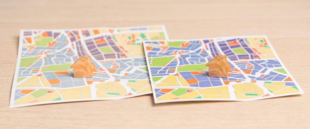 Two printed neighborhood maps with small wooden house figures on a light wood surface.