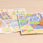 Two printed neighborhood maps with small wooden house figures on a light wood surface.