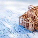 A miniature wooden model of a new construction home frame resting on top of blue architectural blueprints.