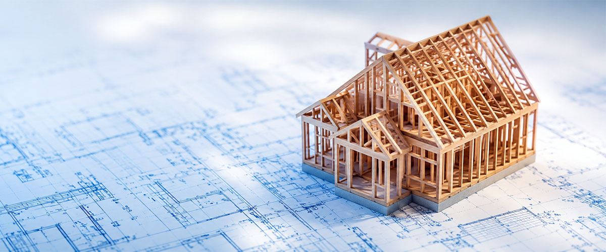 A miniature wooden model of a new construction home frame resting on top of blue architectural blueprints.