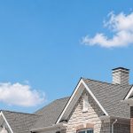 Suburban home rooftops with gray shingles under a bright blue cloudy sky.