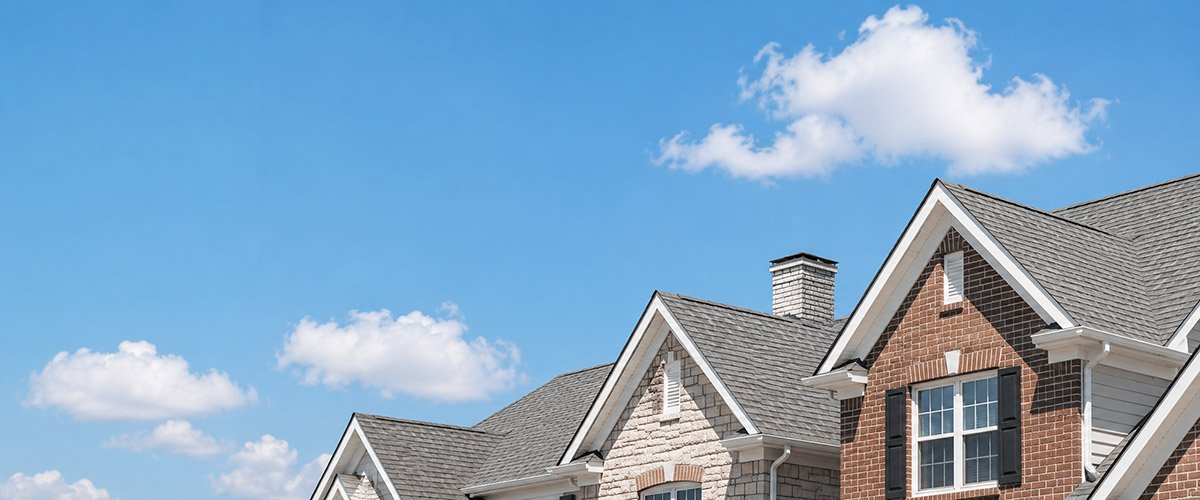 Suburban home rooftops with gray shingles under a bright blue cloudy sky.