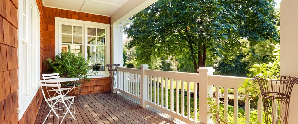Sunlit front porch with white railing, wood decking, bistro table, and leafy yard.