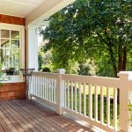 Sunlit front porch with white railing, wood decking, bistro table, and leafy yard.