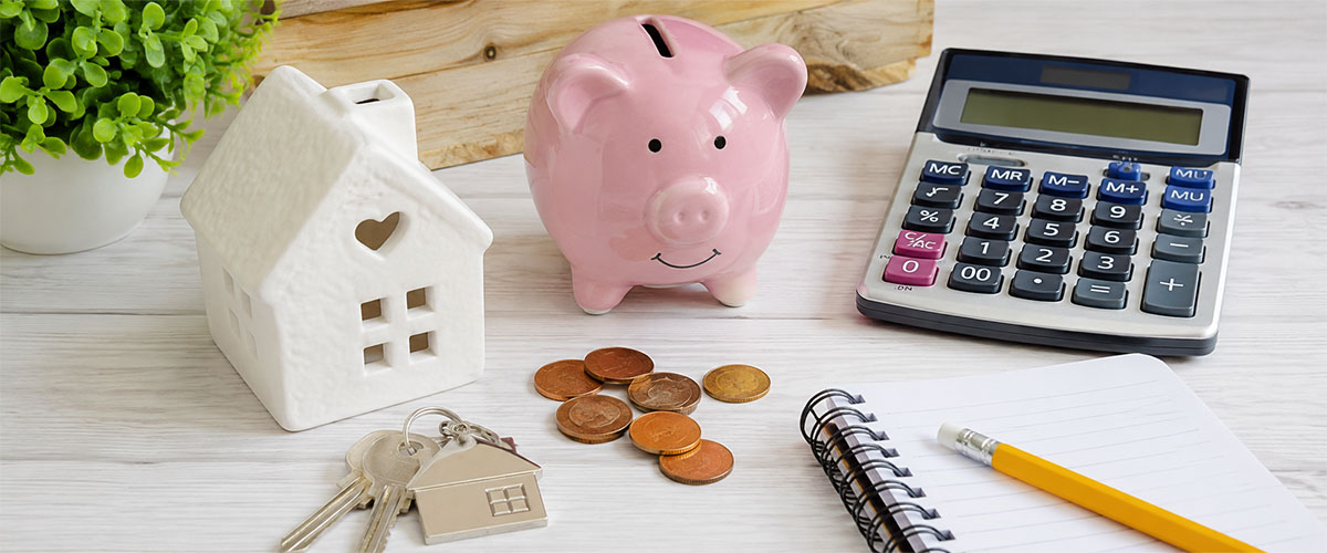White desk displaying a ceramic house, pink piggy bank, calculator, coins, and keys.