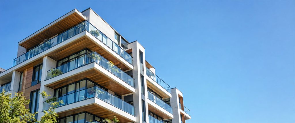 Contemporary condominium building with glass-railed balconies, white concrete, and warm wood accents beneath a clear blue sky.
