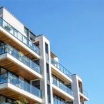 Contemporary condominium building with glass-railed balconies, white concrete, and warm wood accents beneath a clear blue sky.