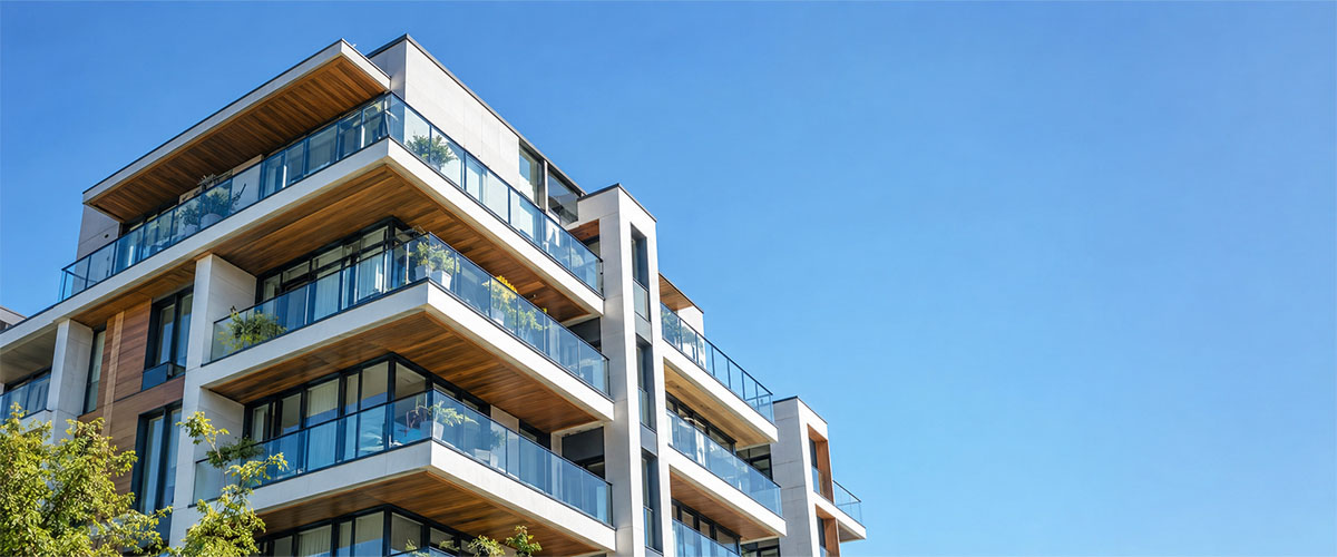 Contemporary condominium building with glass-railed balconies, white concrete, and warm wood accents beneath a clear blue sky.