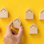 Hand placing a wooden house block among a group to represent available housing inventory.