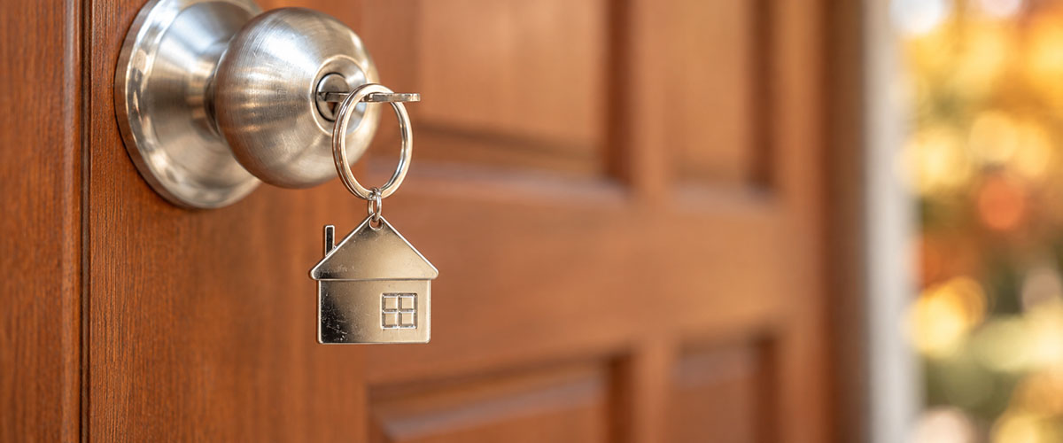 House-shaped keychain hangs from a key in a silver door lock beside blurred fall foliage.