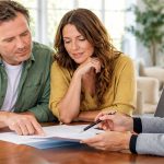 Three people review paperwork together at a table with a laptop nearby.