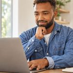 Man in a denim shirt using a laptop at a table beside a bright window.