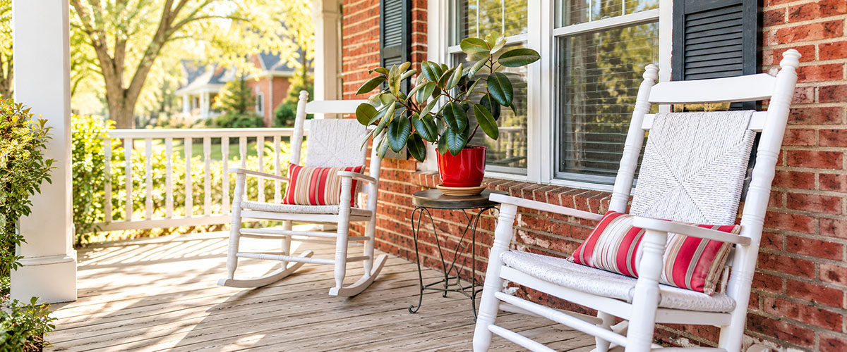 White rocking chairs and a potted plant sit on a wooden front porch.