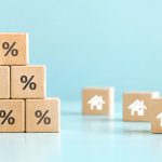 Wooden blocks with percent symbols stacked beside house icons on a pale blue background, representing mortgage rates.