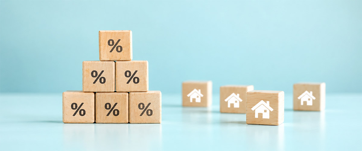 Wooden blocks with percent symbols stacked beside house icons on a pale blue background, representing mortgage rates.